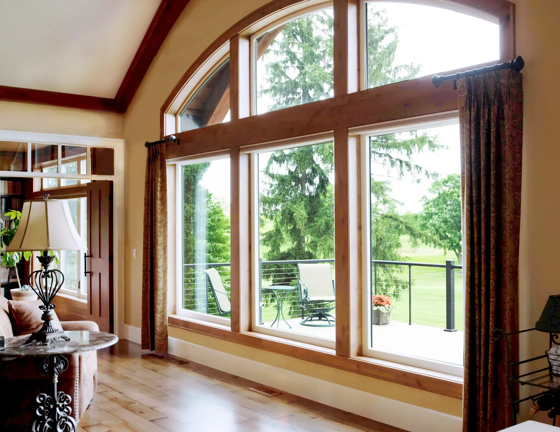Spacious living room with large wood-framed windows and glass doors overlooking a deck and forested landscape, featuring cream walls, wooden beams, and burgundy curtains