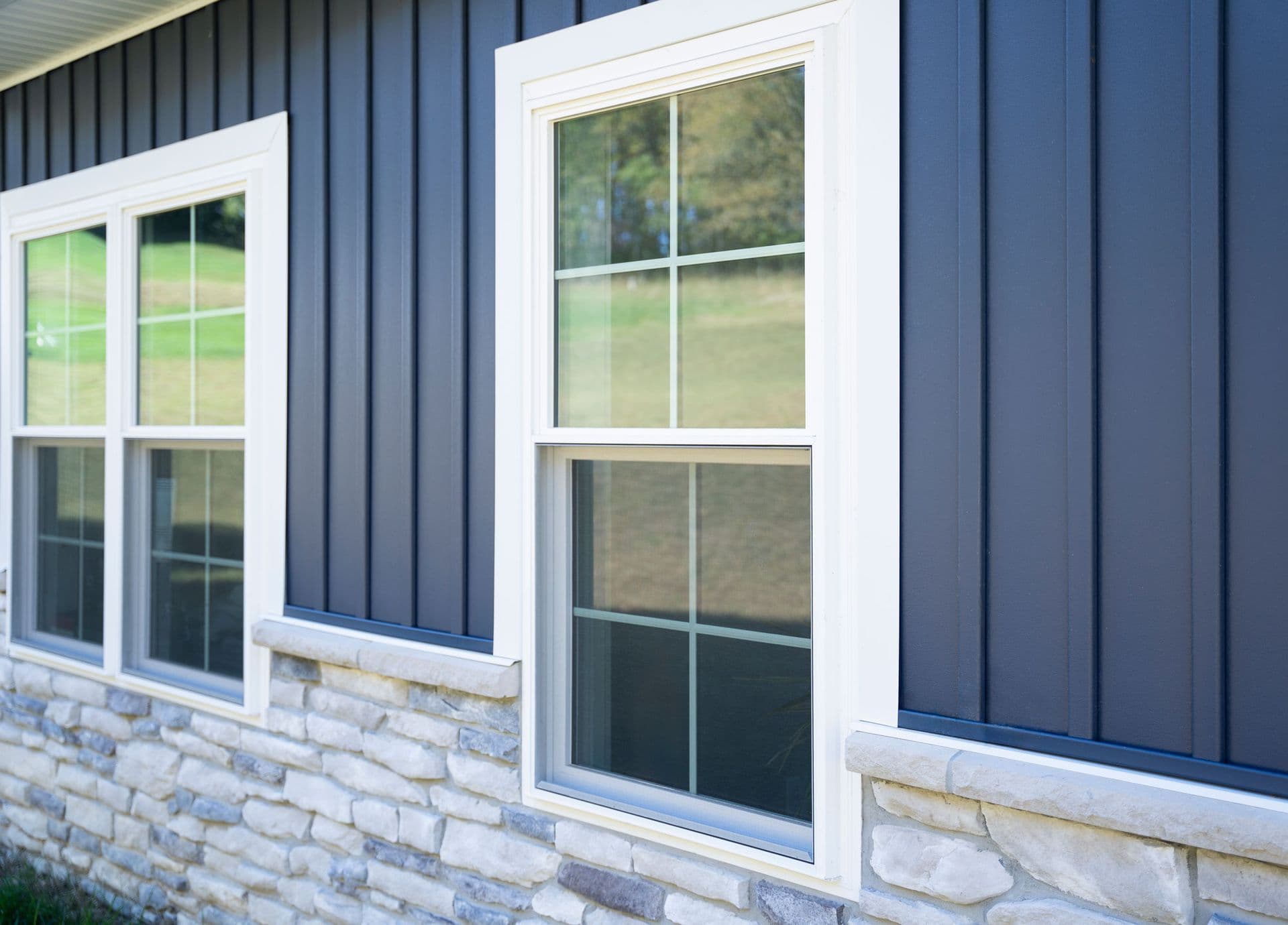 Modern building exterior with white-framed windows and blue corrugated metal siding above stone foundation, showing landscaped grounds through glass panes