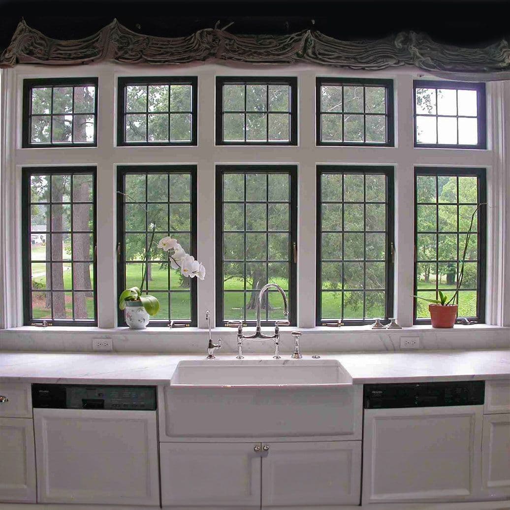 Kitchen sink with farmhouse apron basin and chrome faucet beneath a grid of black-framed windows overlooking gardens and trees, with potted plants on the windowsill
