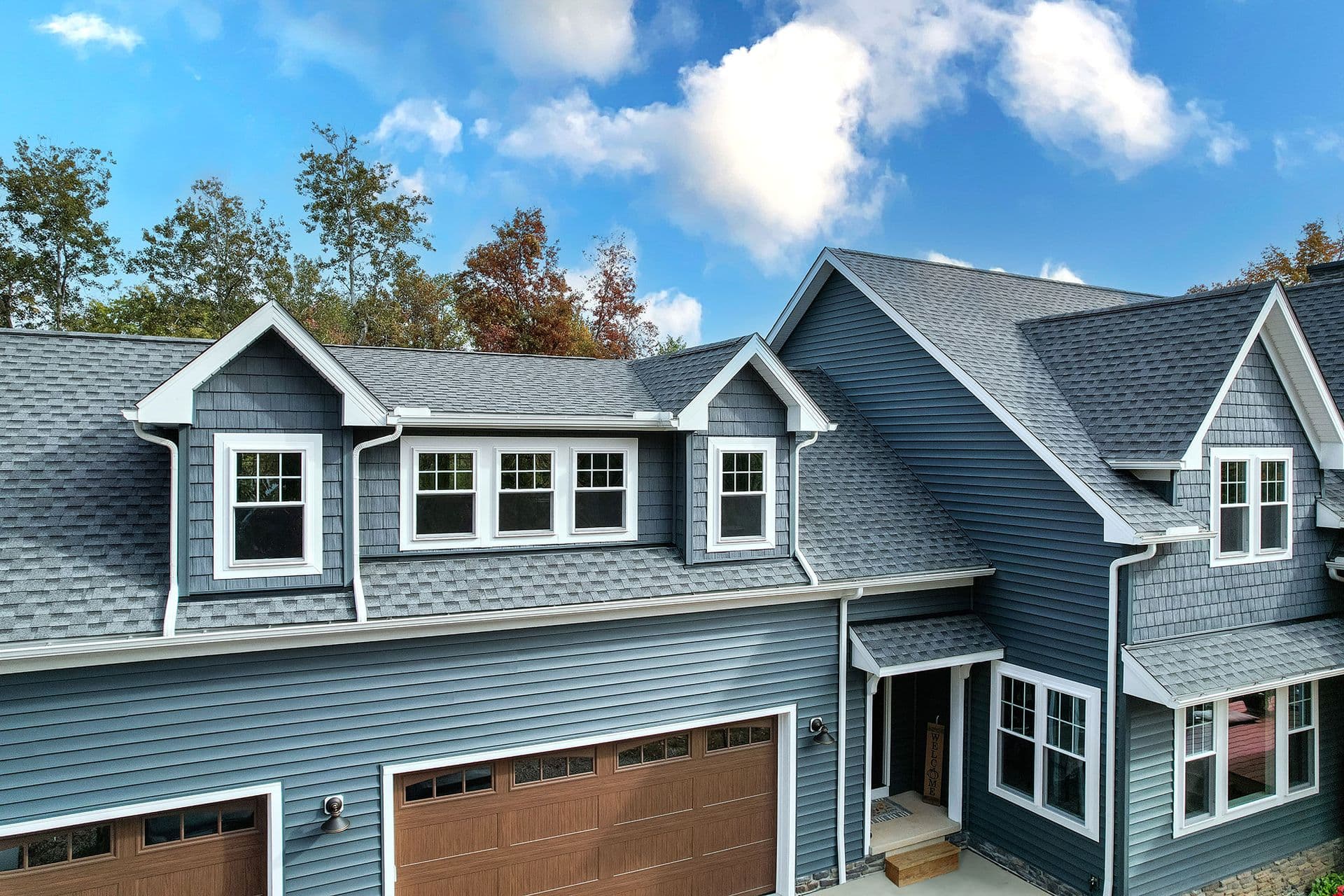 Modern two-story home with blue-gray siding, white trim, multiple gabled rooflines, dormer windows, and attached two-car garage with wood-panel doors, surrounded by autumn trees