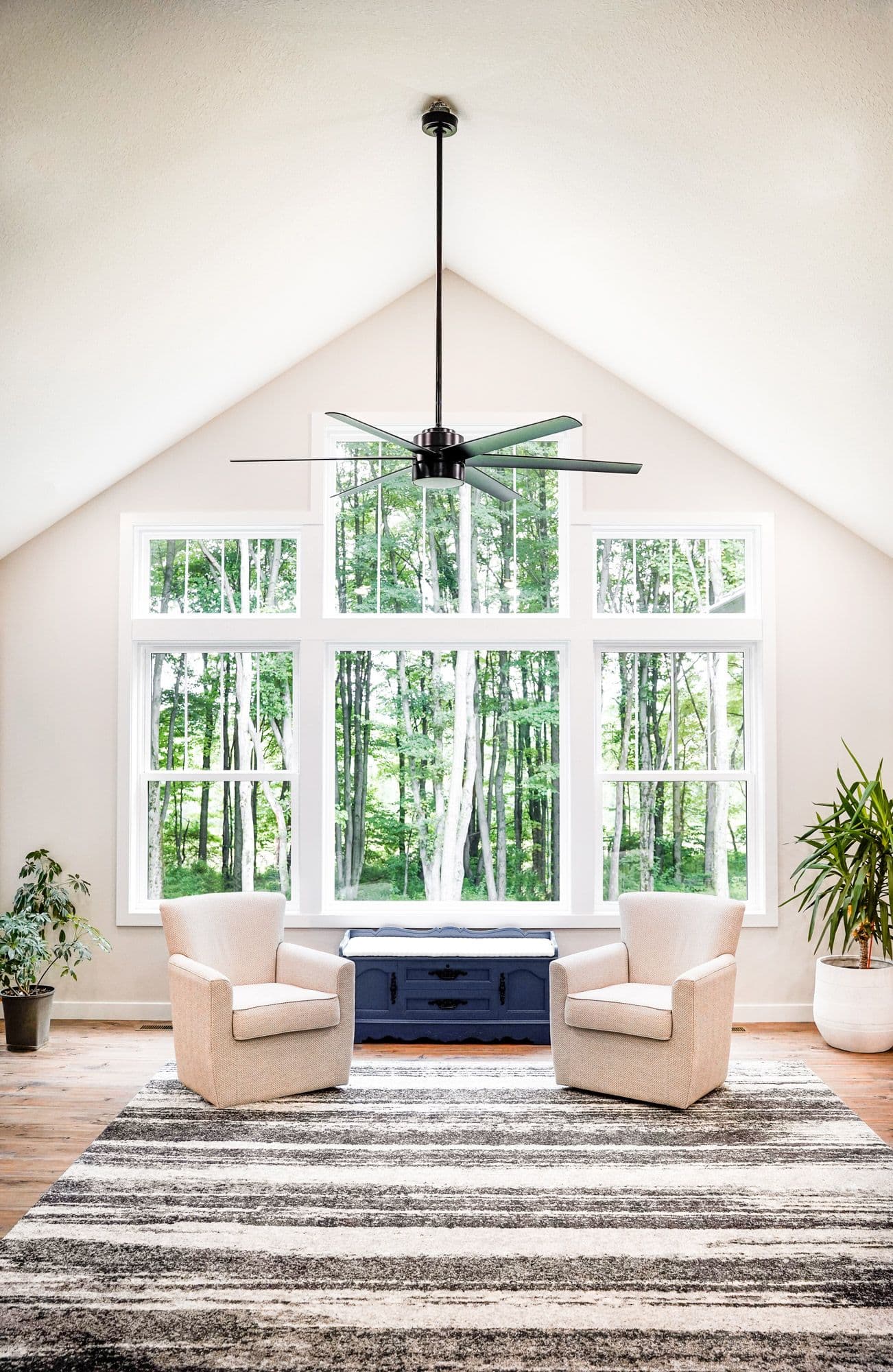 Modern living room with vaulted ceiling, black ceiling fan, two cream armchairs, navy storage bench, and large arched window overlooking forest trees